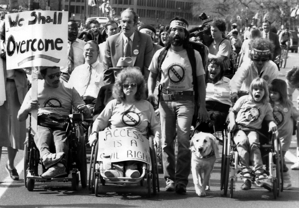 man in wheelchair with We Shall Overcome sign over his head rolling next to woman in wheelchair, next to blind guy with dog guide next to young girl in wheelchair. They are in front of crowd of other marchers