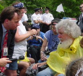 Mark McClellan, in suit with White House badge around his neck leans over to talk with Bob Kafka, in yellow rain poncho. All around are other ADAPTers and police stand in the background.